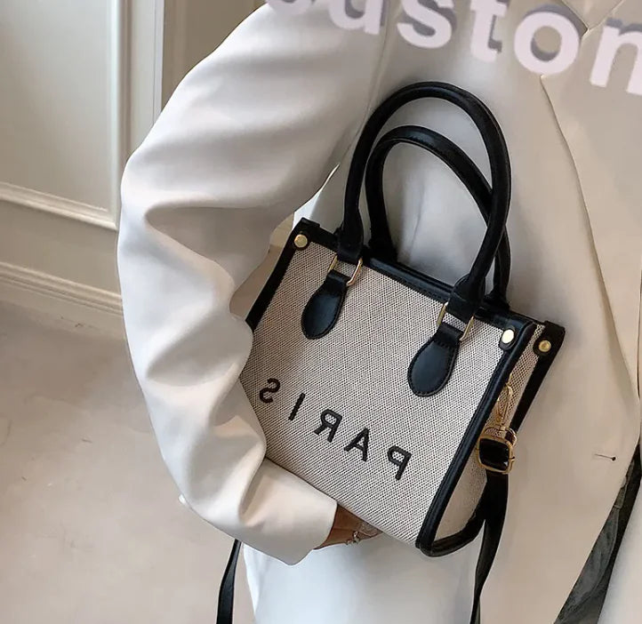 Person holding a beige handbag with black handles and 'PARIS' text, against a white background
